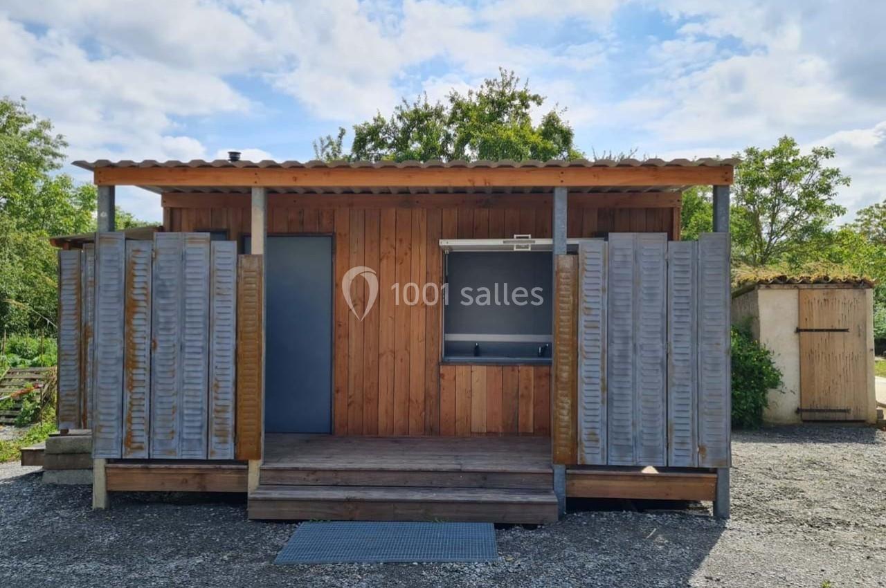 Cabane en bois avec volets gris, une porte et un comptoir ouvert, située sur un terrain extérieur gravillonné.