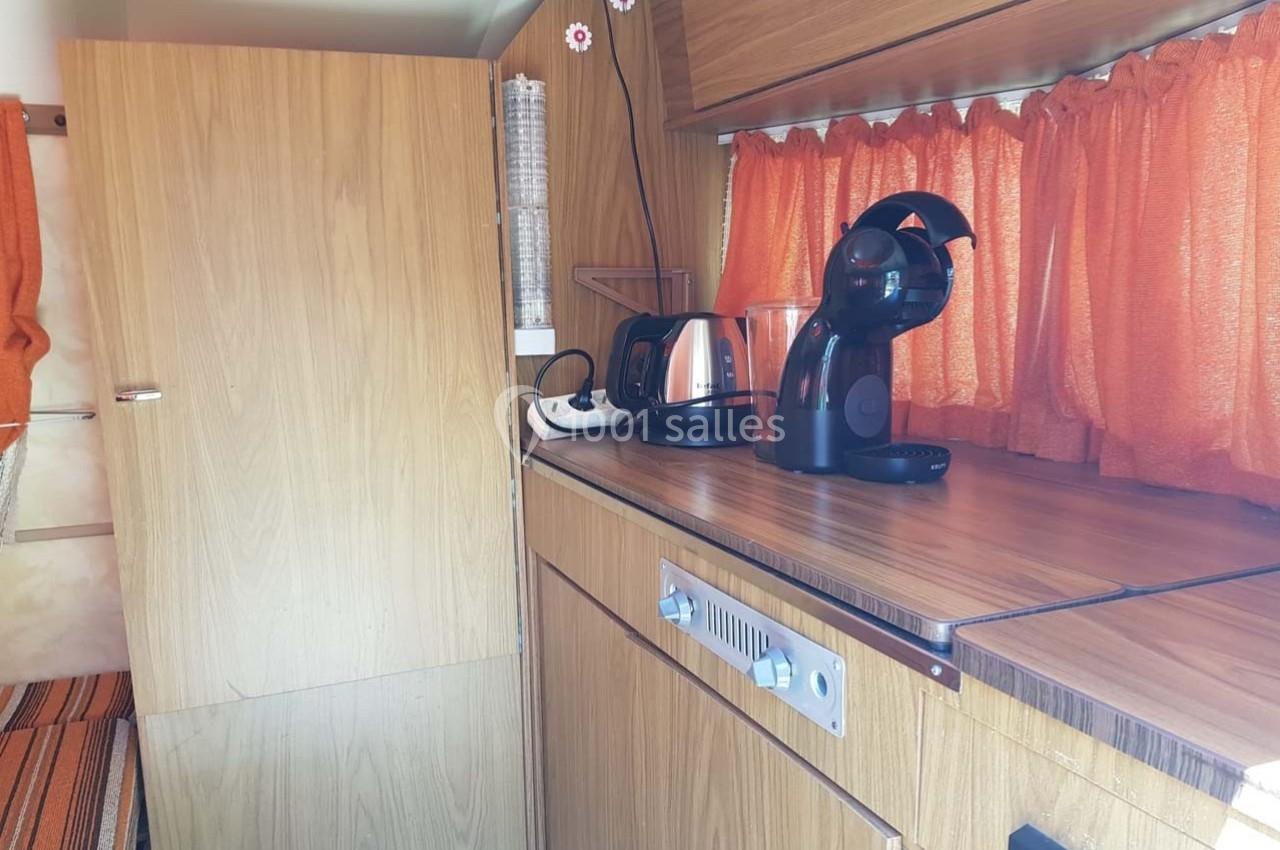 Intérieur d'un camping-car avec un plan de travail en bois, une cafetière, une bouilloire et des rideaux orange.