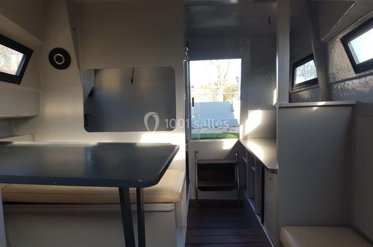 Intérieur d'un petit espace de vie avec table, banquettes, fenêtres et vue sur l'extérieur par une porte ouverte.