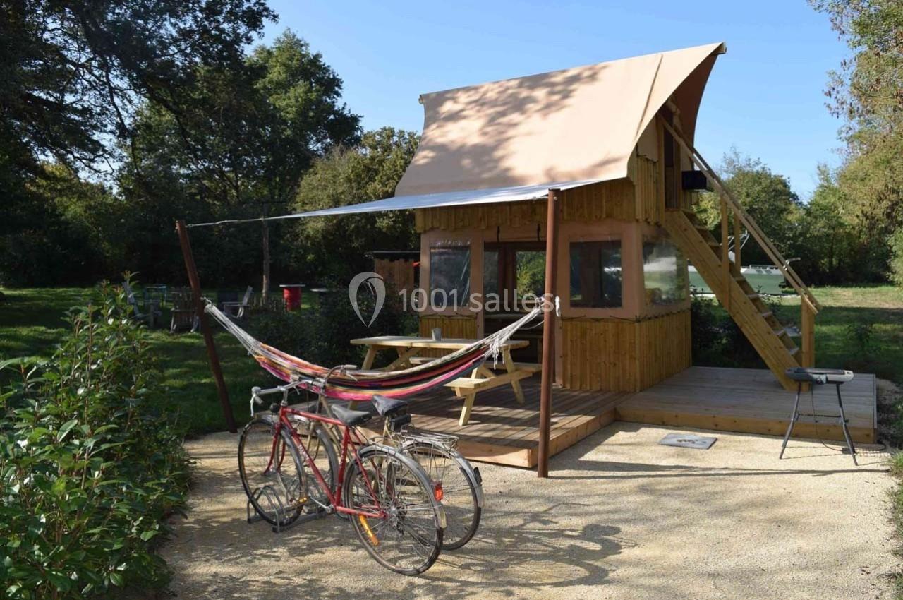 Cabane en bois avec terrasse, hamac, vélos et table de pique-nique, située dans un environnement naturel verdoyant.