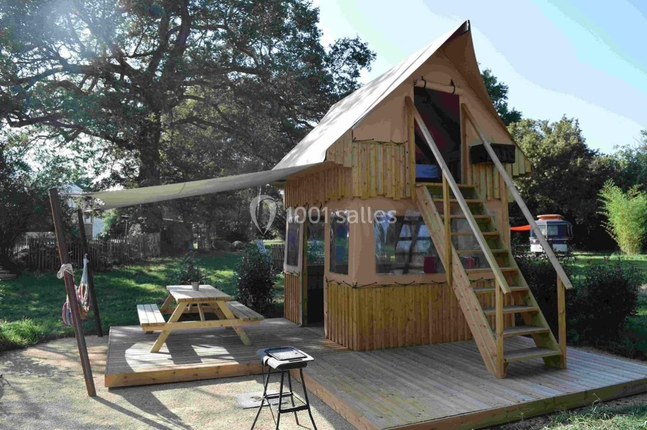 Cabane en bois surélevée avec terrasse, table de pique-nique, auvent et escalier, située dans un jardin arboré.