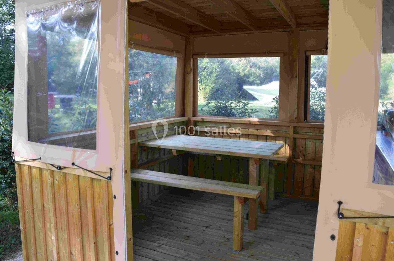 Abri en bois avec table et bancs intégrés, entouré de fenêtres transparentes donnant sur un espace extérieur verdoyant.