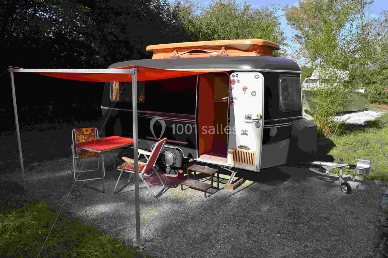 Caravane vintage noire avec auvent orange, table et chaise installées sur un emplacement extérieur ombragé.