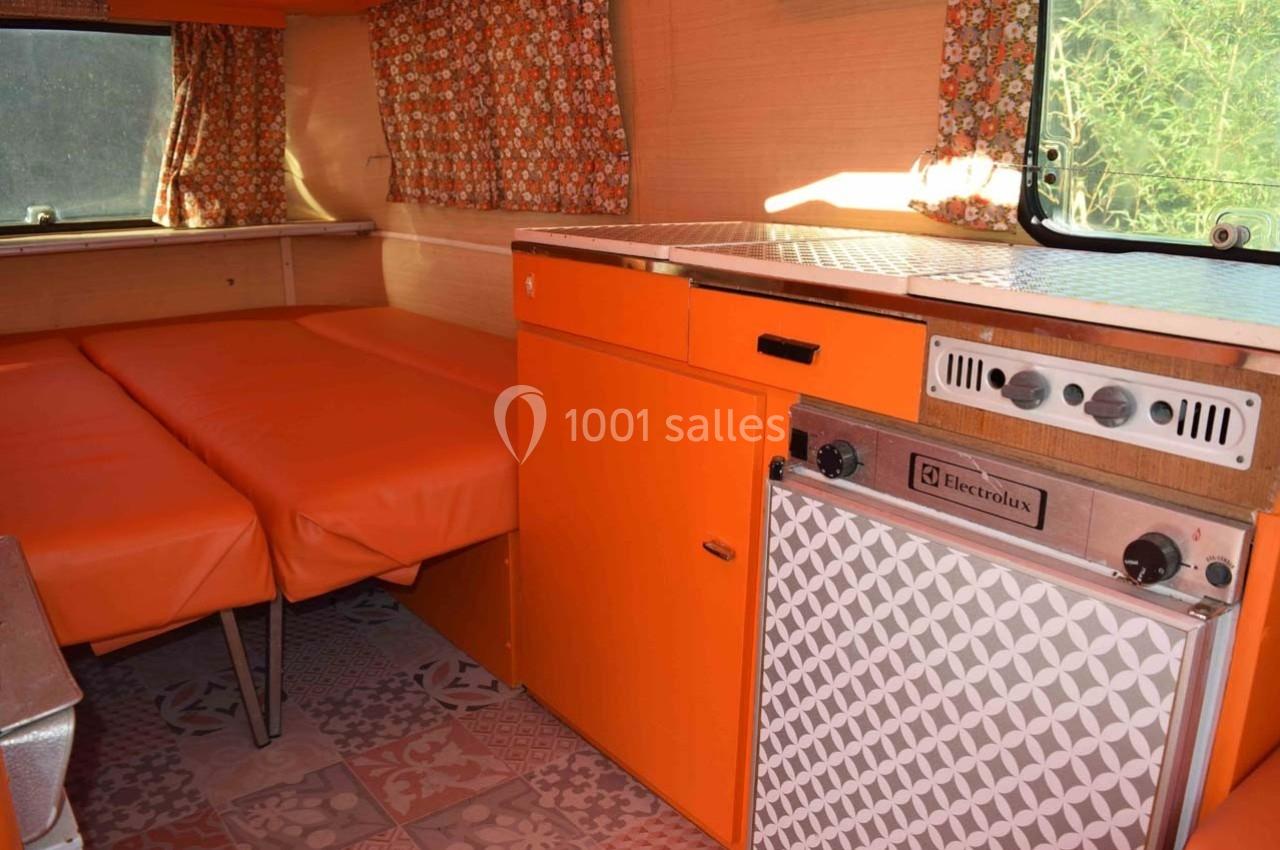 Intérieur d'une caravane vintage avec banquettes orange, rideaux à motifs et kitchenette équipée d'un réfrigérateur.