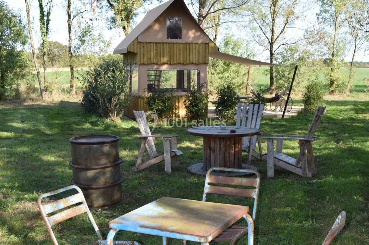Mobilier de jardin rustique avec chaises, tables et tonneaux devant une petite cabane en bois dans un cadre verdoyant.