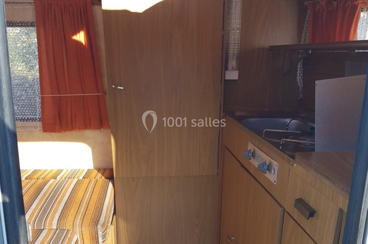 Intérieur d'une caravane avec un coin cuisine en bois, un évier, des rideaux orange et un espace couchage à l'arrière.