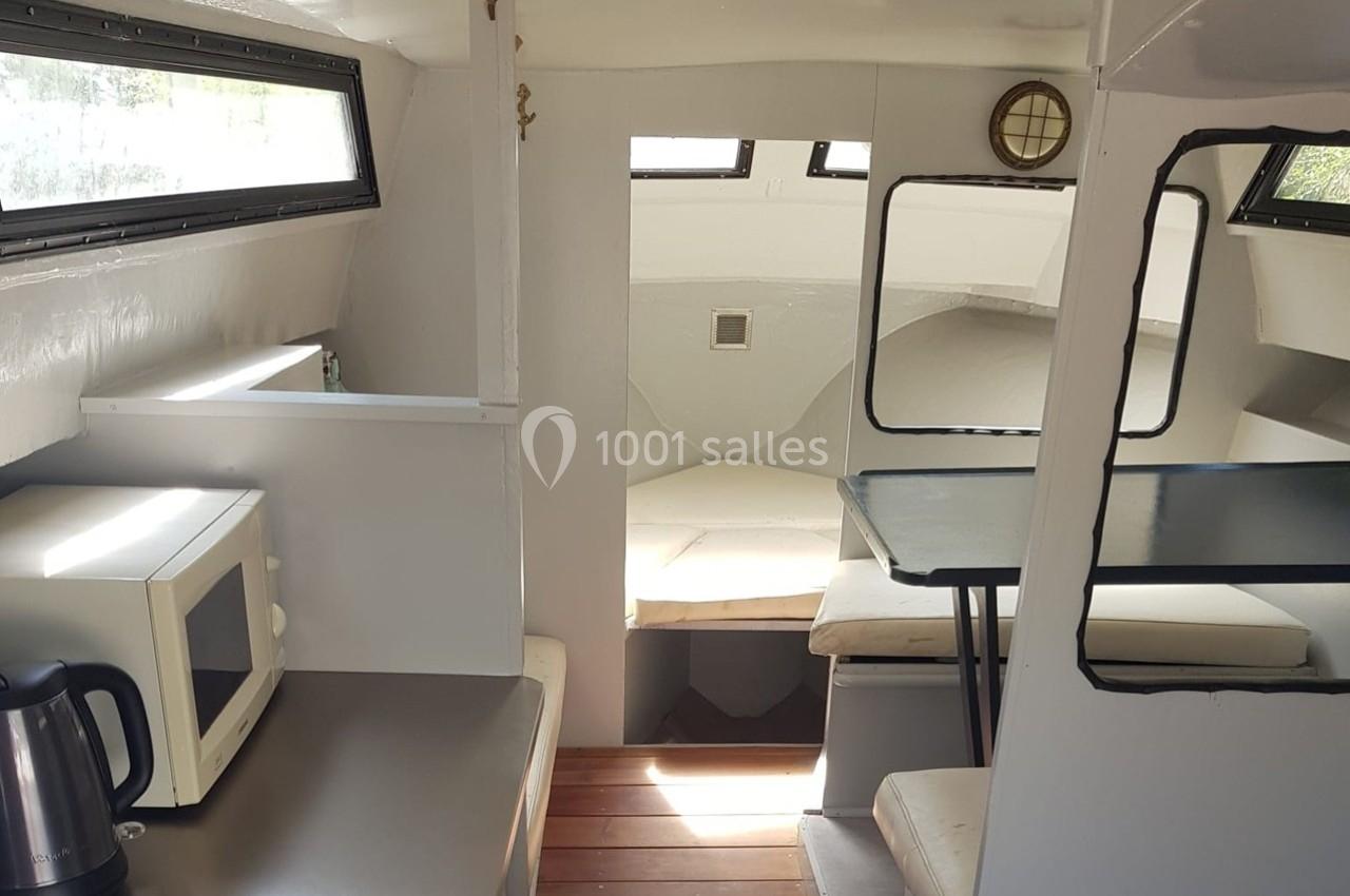 Intérieur d'un petit bateau avec coin cuisine équipé d'un micro-ondes, d'une bouilloire et espace repas avec banquettes.