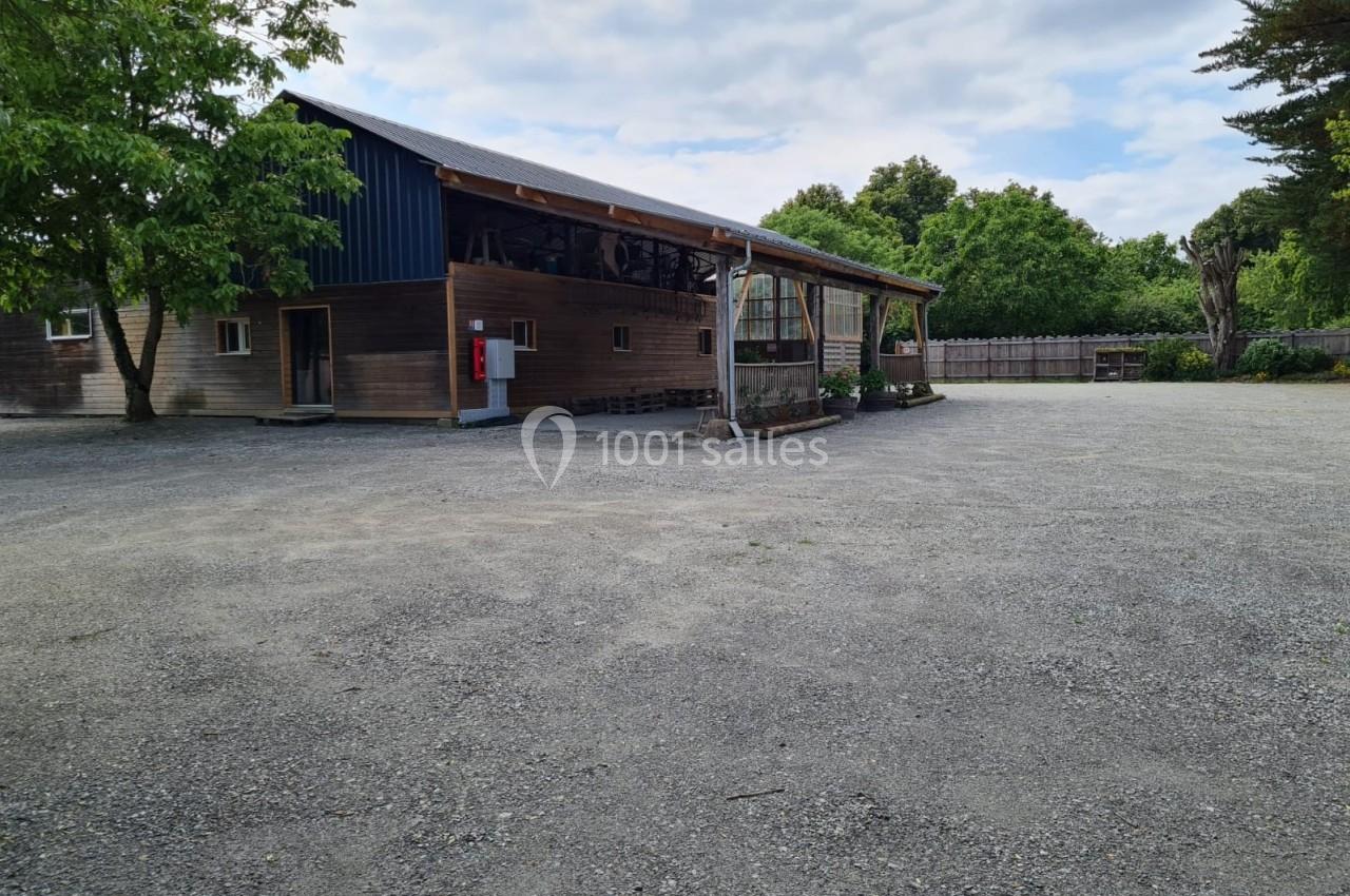 Bâtiment en bois avec toit en tôle, entouré d'arbres, donnant sur une grande cour en gravier.