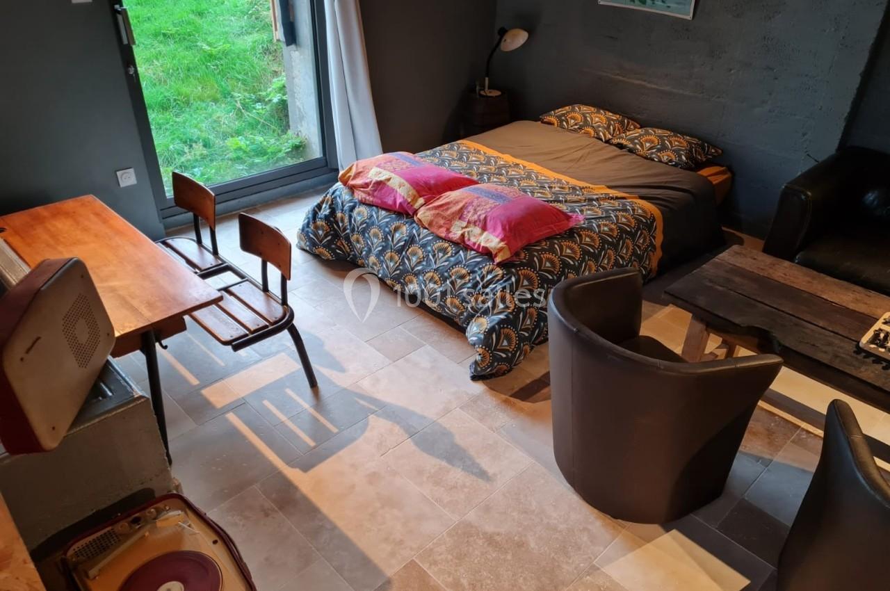 Chambre avec lit double, bureau, chaises, fauteuils, tourne-disque et grande fenêtre donnant sur un jardin.