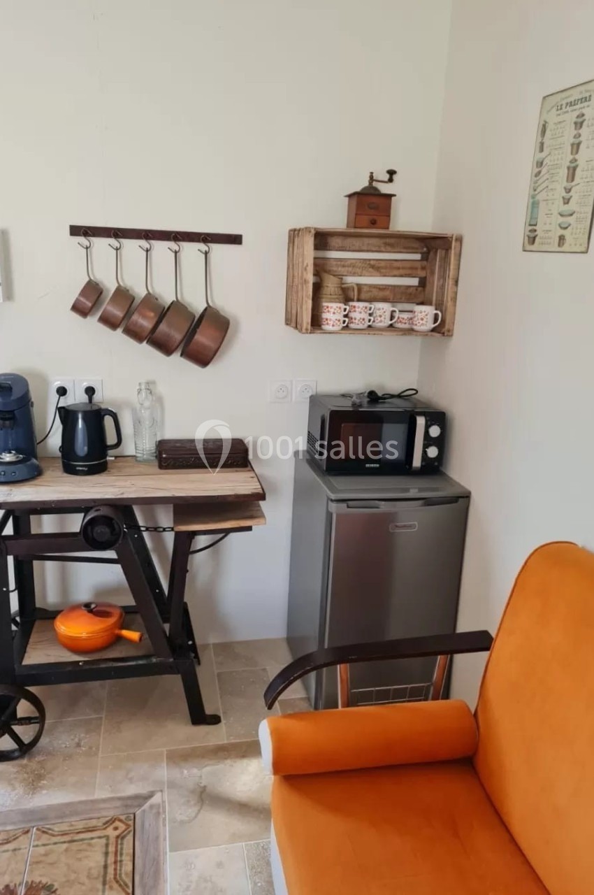 Coin cuisine avec cafetière, bouilloire, micro-ondes, casseroles suspendues, étagère en bois et fauteuil orange au premier…