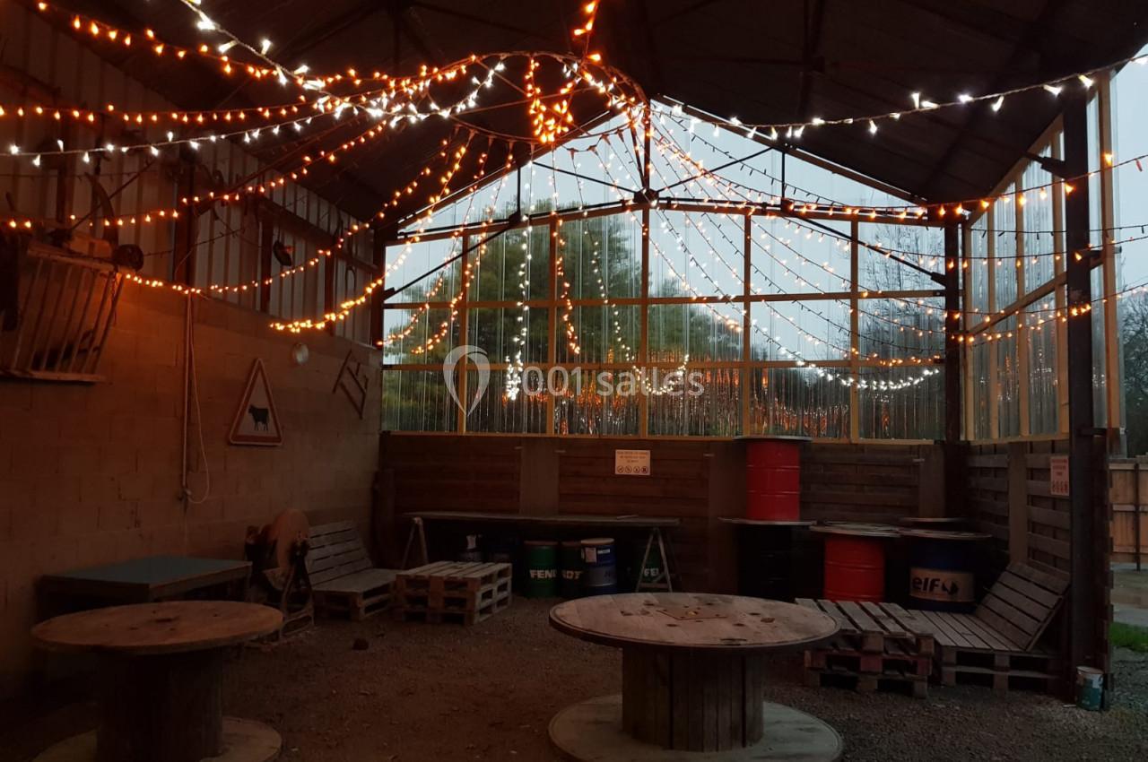 Espace couvert avec guirlandes lumineuses, tables en bois et murs en palettes, ambiance rustique et chaleureuse.