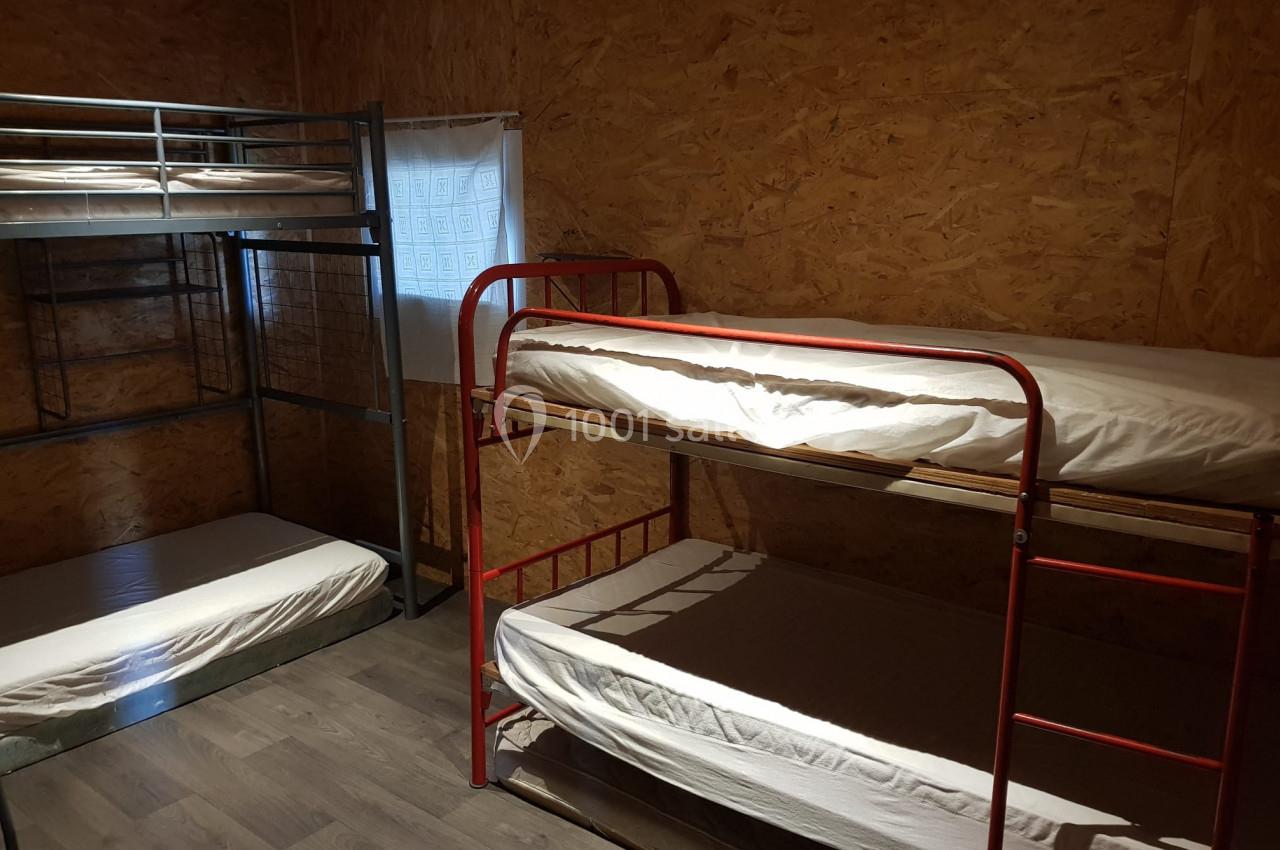 Chambre simple avec lits superposés en métal, murs en bois et fenêtre avec rideau blanc.