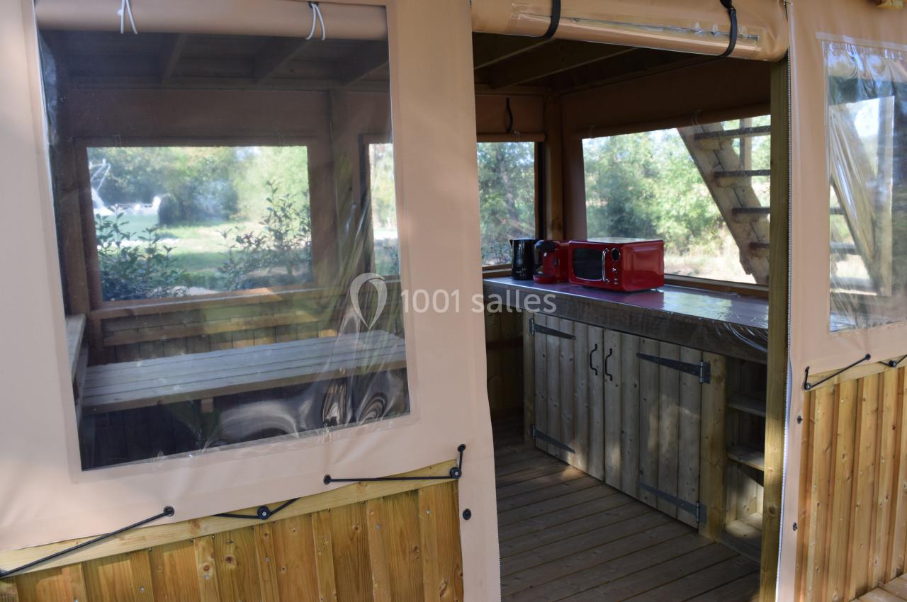 Intérieur d'une tente en bois avec une table, des bancs et un plan de travail équipé de petits appareils électroménagers.