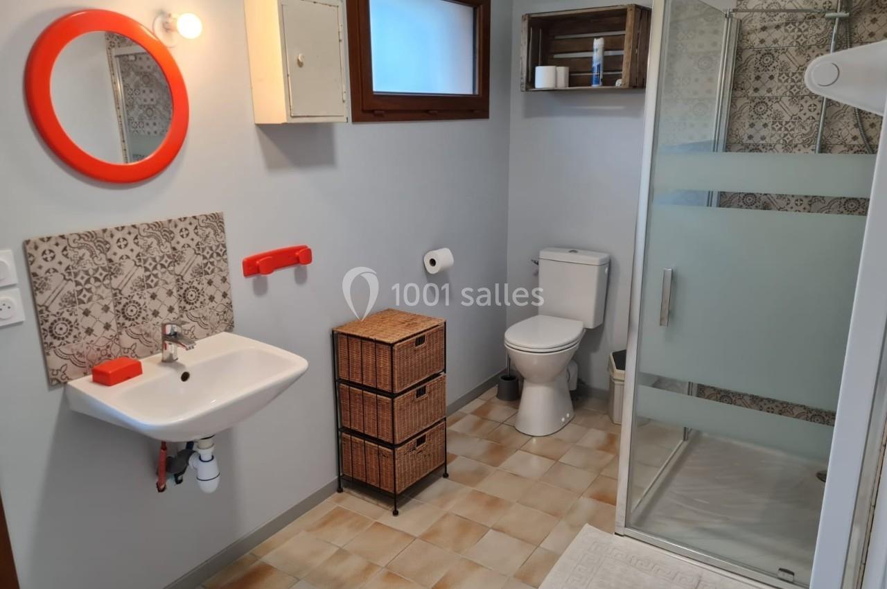 Salle de bain avec lavabo, miroir rond rouge, douche vitrée, toilettes et meubles de rangement en osier.