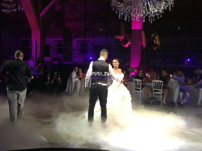 Un couple danse sous un éclairage coloré dans une salle décorée, entouré d'invités assis et d'une ambiance de fumée au sol.