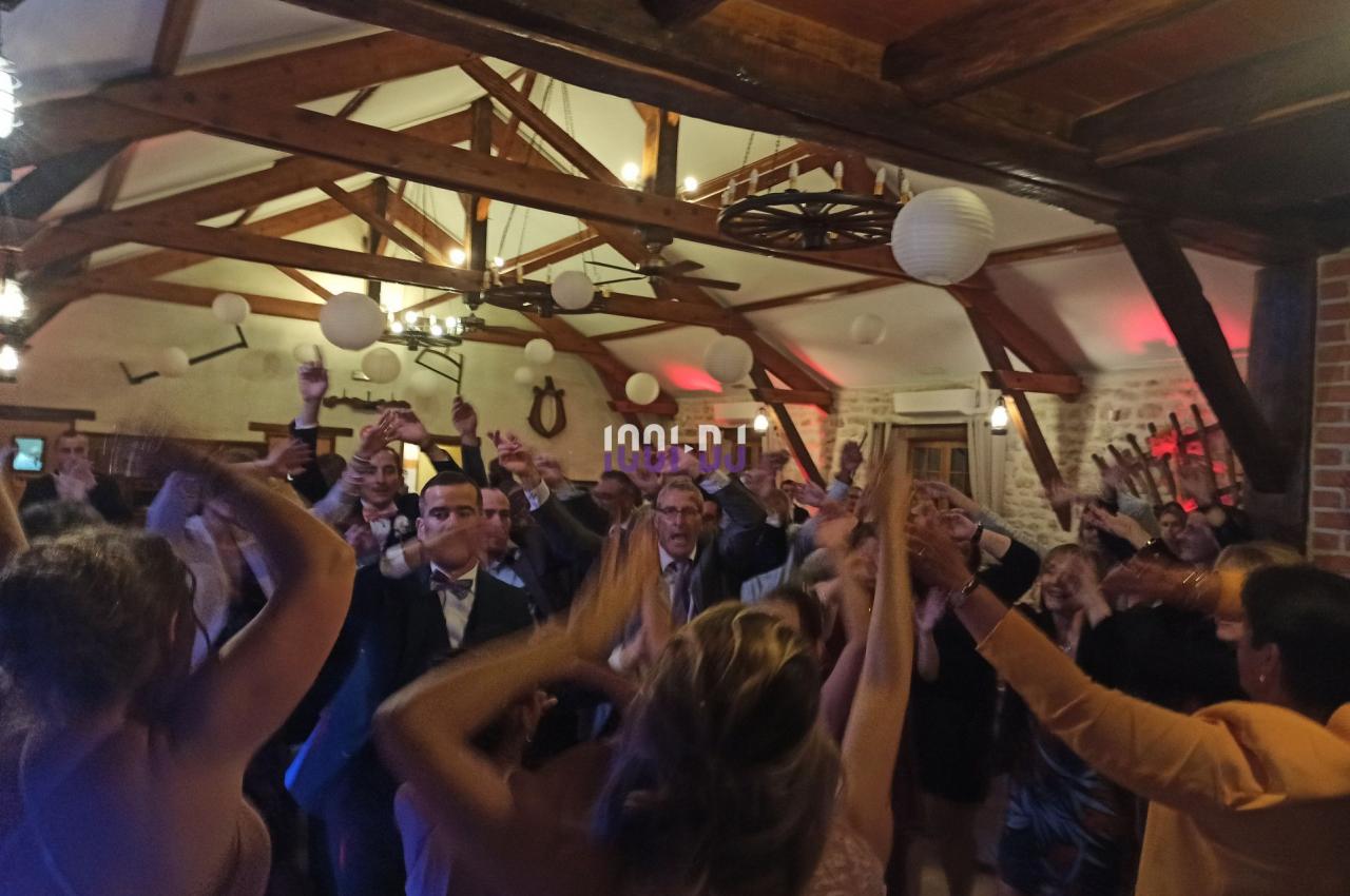 Des personnes dansent et lèvent les bras lors d'une fête dans une salle avec poutres apparentes et décorations suspendues.