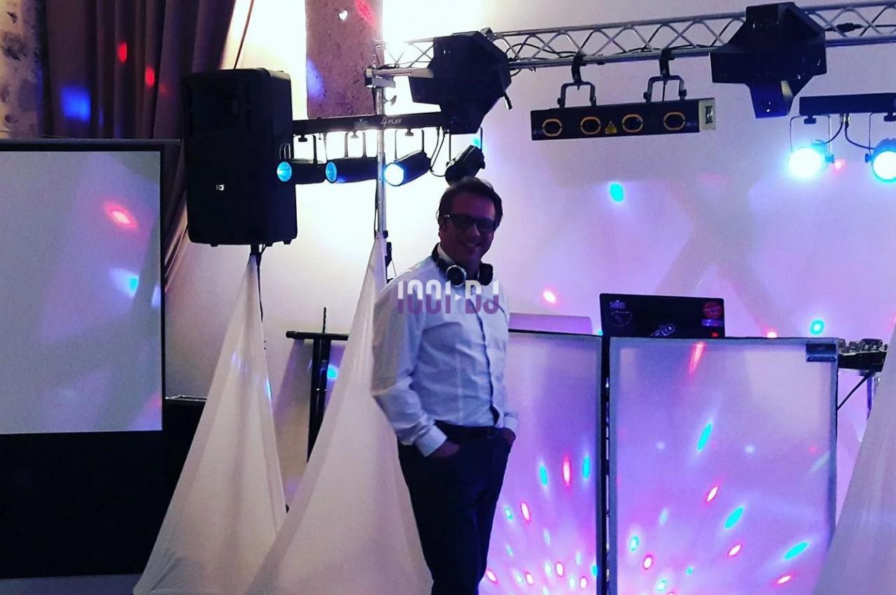 Un DJ souriant se tient devant son équipement lumineux dans une salle décorée pour un événement.