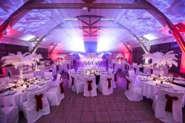 Salle de réception décorée avec des tables rondes, nappes blanches, nœuds rouges et éclairage coloré.