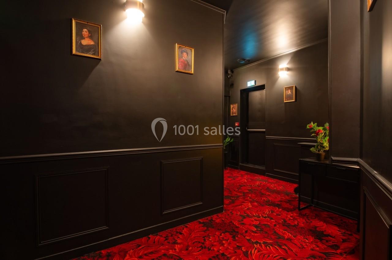 Couloir sombre avec murs noirs, tapis rouge à motifs, tableaux encadrés et petite table décorée de fleurs.