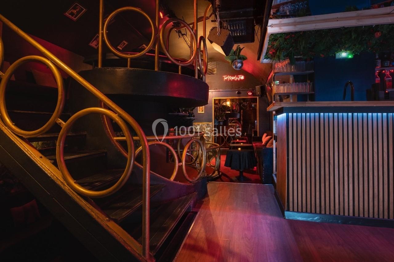 Escalier en colimaçon noir avec rampes dorées, bar éclairé en bleu et rouge, décor végétal et ambiance tamisée.