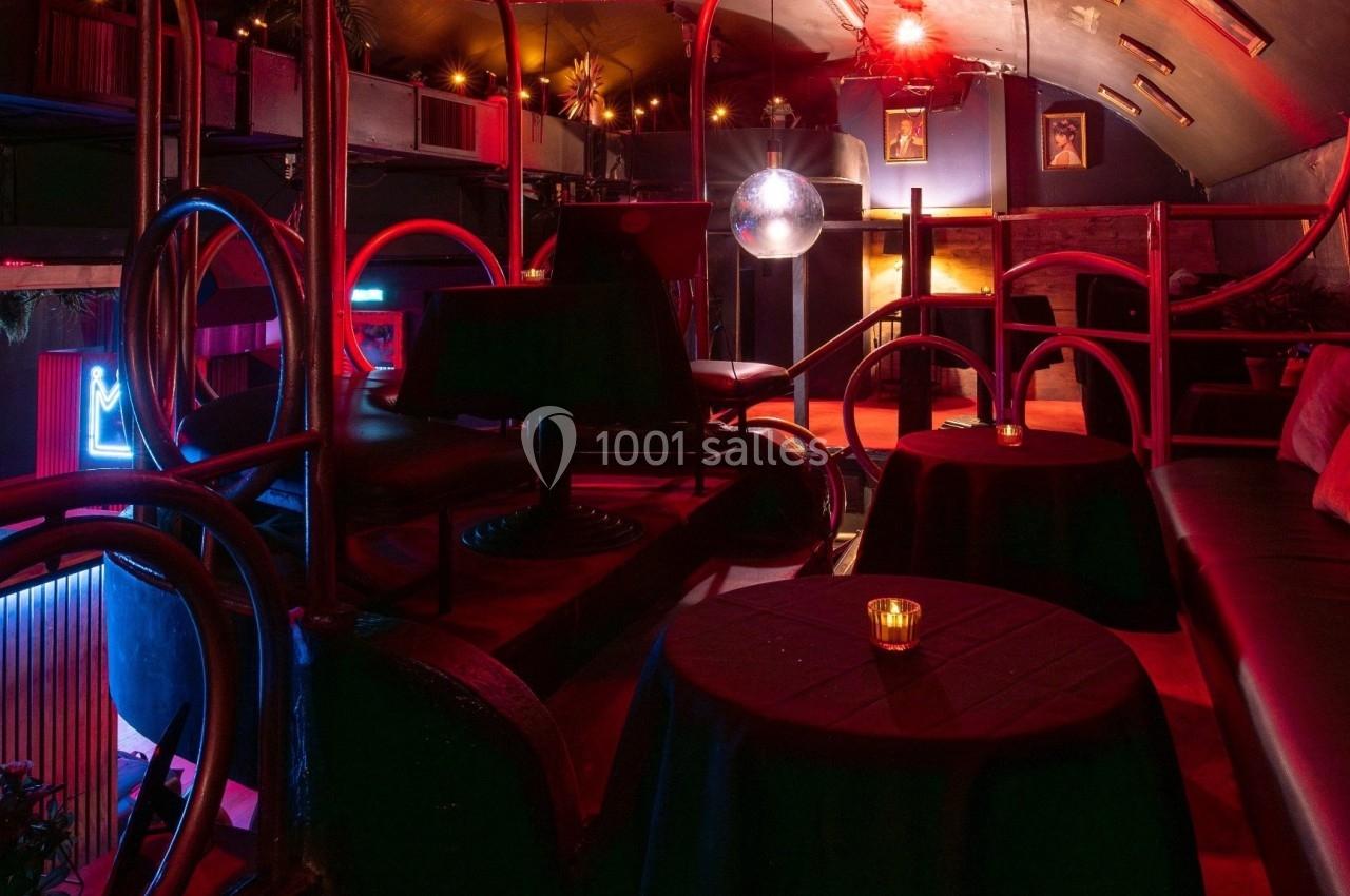 Salle intimiste avec éclairage tamisé rouge, tables rondes avec bougies, banquettes et décorations rétro.