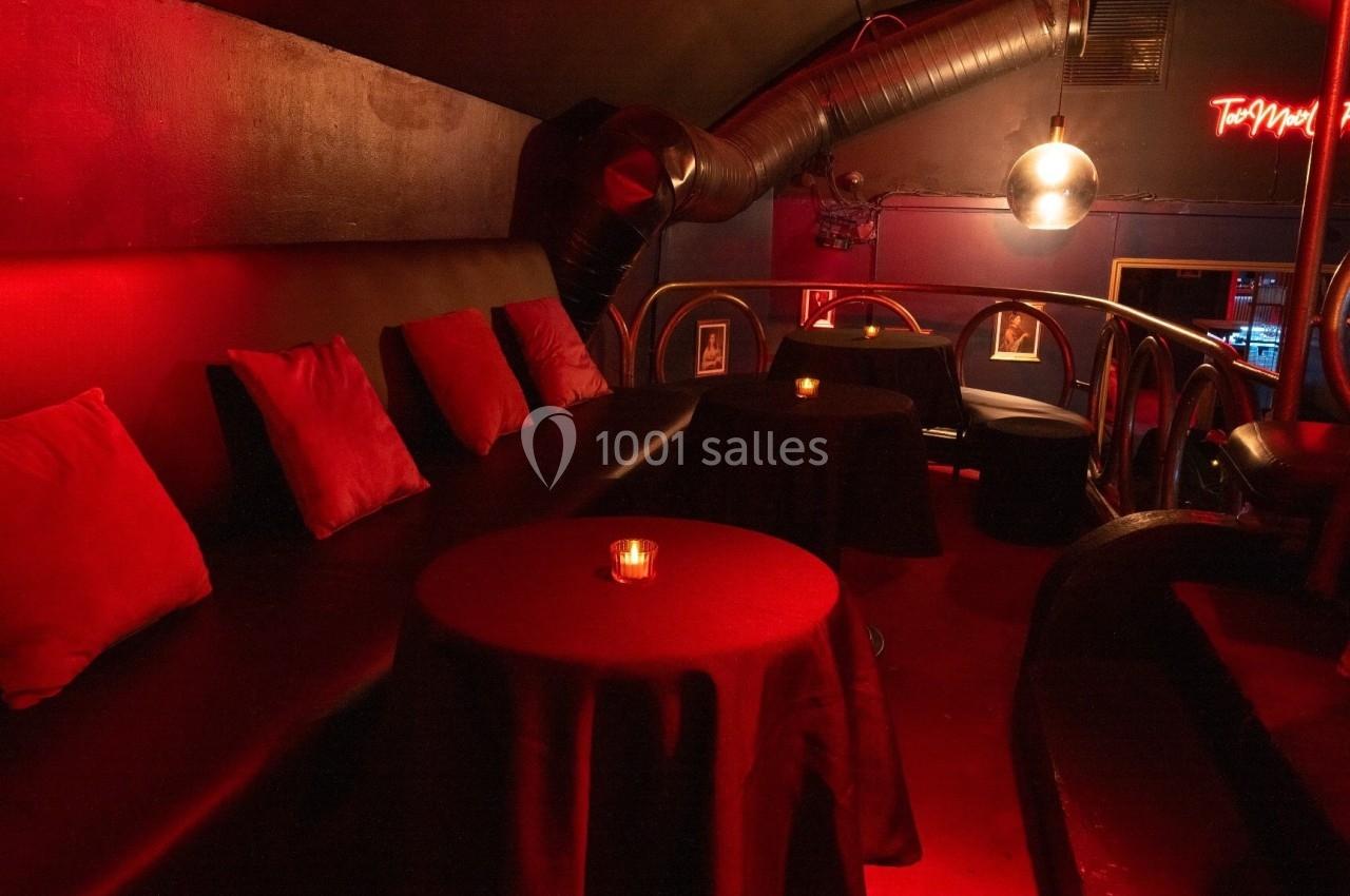 Espace lounge sombre avec banquettes noires, coussins rouges, tables rondes et éclairage tamisé.