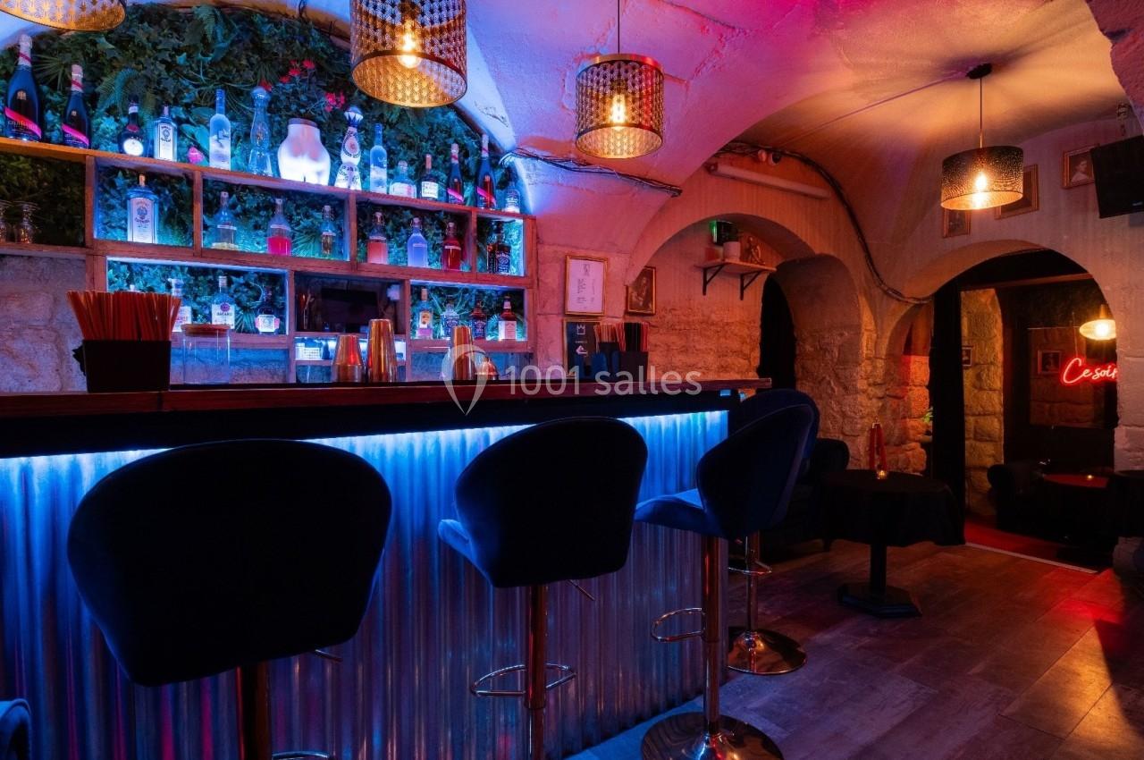 Bar avec comptoir éclairé, tabourets hauts, étagères de bouteilles illuminées et ambiance tamisée dans une cave voûtée.