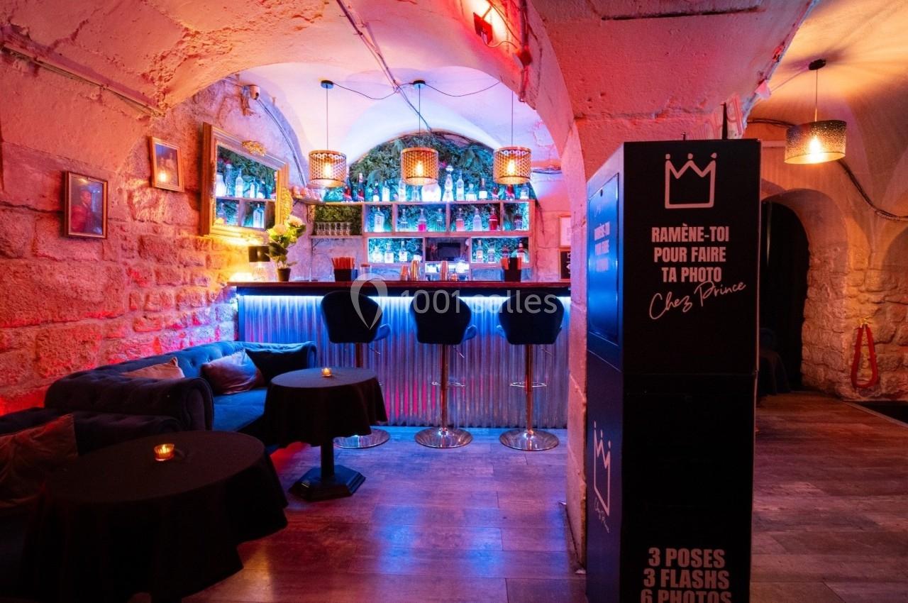 Intérieur d'un bar avec un comptoir éclairé, des tabourets hauts, des tables basses et une borne photo.