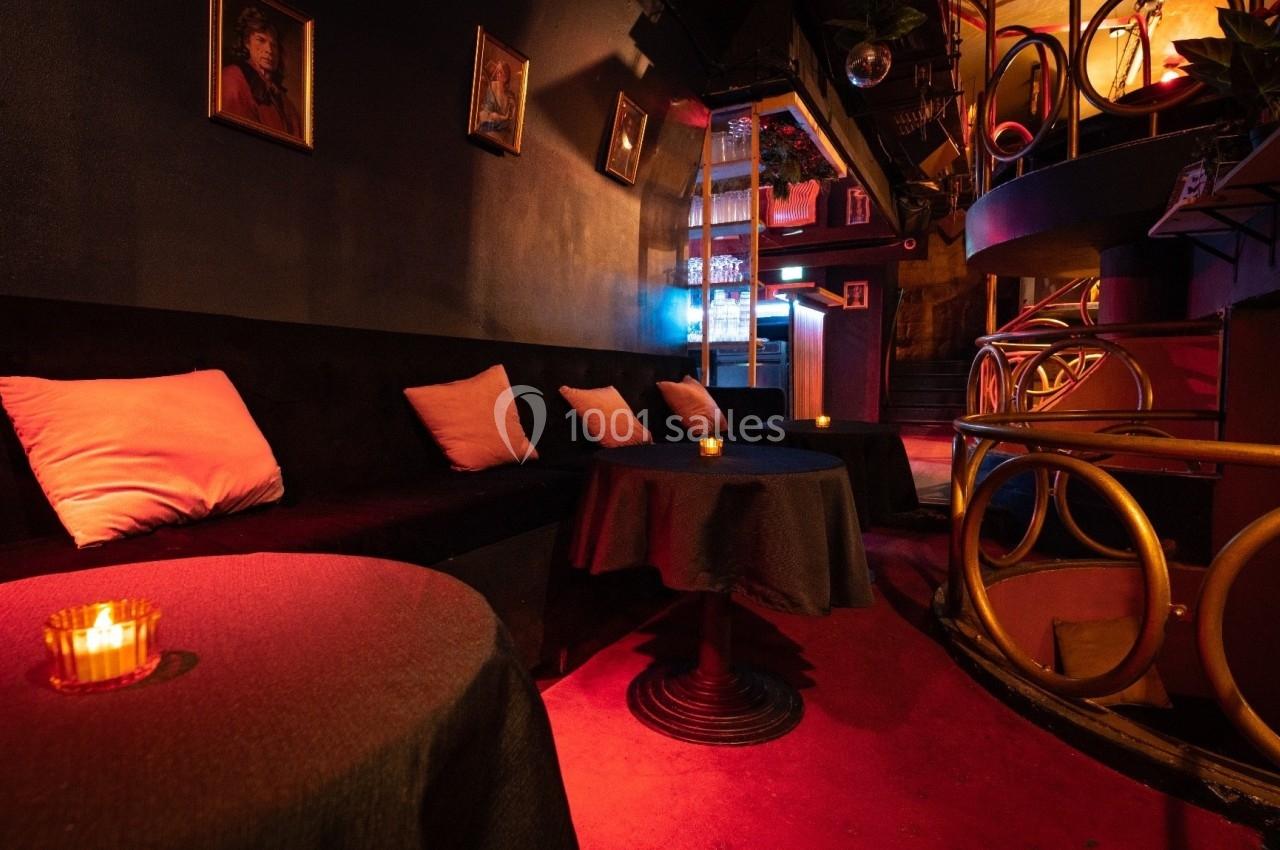 Salle sombre avec tables rondes, bougies allumées, banquettes noires et coussins, décorée de cadres au mur.