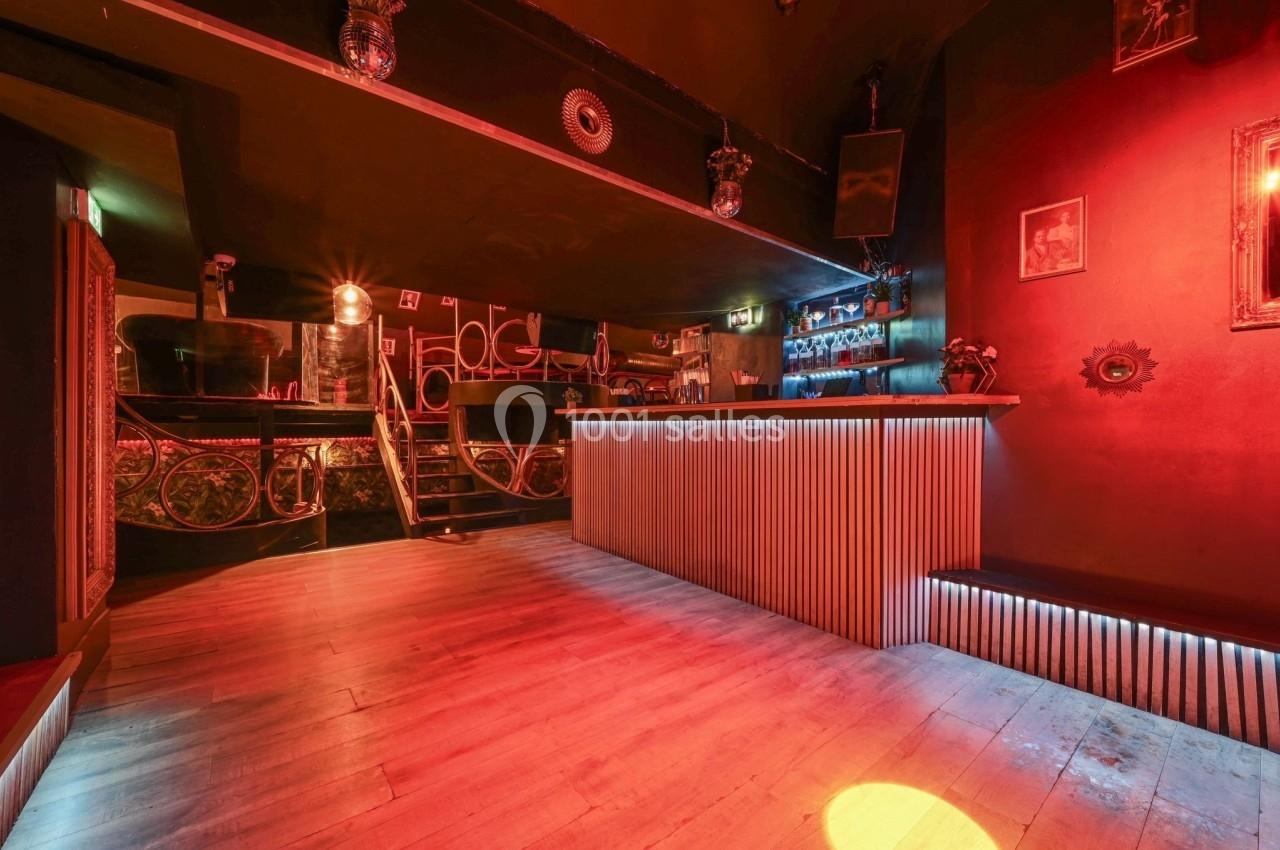 Intérieur d'un bar avec un comptoir éclairé, un sol en bois et une ambiance lumineuse rouge tamisée.