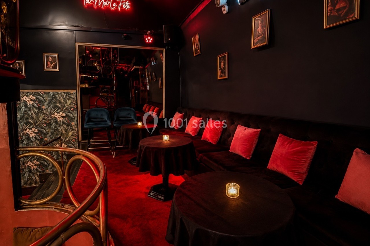 Salle sombre et cosy avec banquettes noires, coussins rouges, tables rondes et bougies, décorée de cadres muraux.