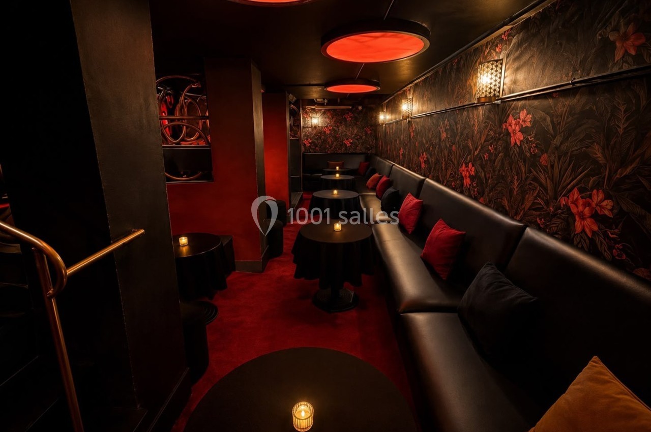 Salle intimiste avec banquettes noires, tables rondes, éclairage tamisé rouge et murs décorés de motifs floraux.