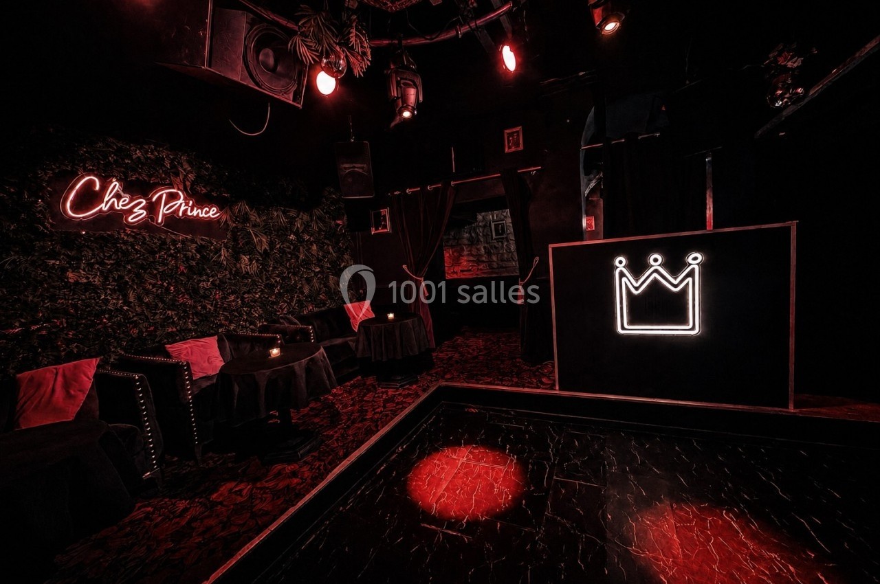 Salle sombre avec éclairage rouge, tables basses, fauteuils, enseigne ’Chez Prince’ et logo de couronne lumineux.