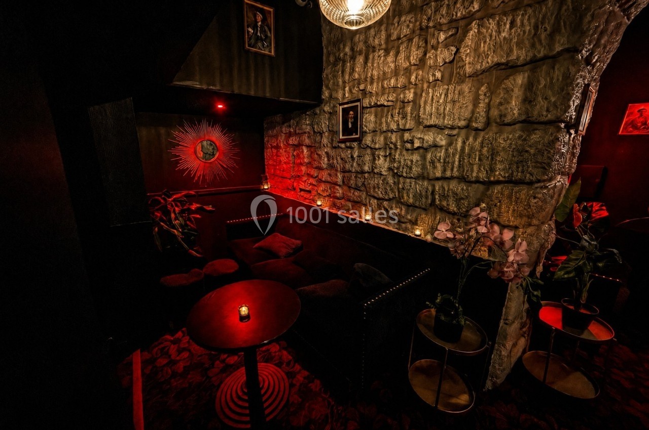 Coin salon sombre avec canapé noir, murs en pierre, éclairage rouge tamisé et petite table avec bougie.