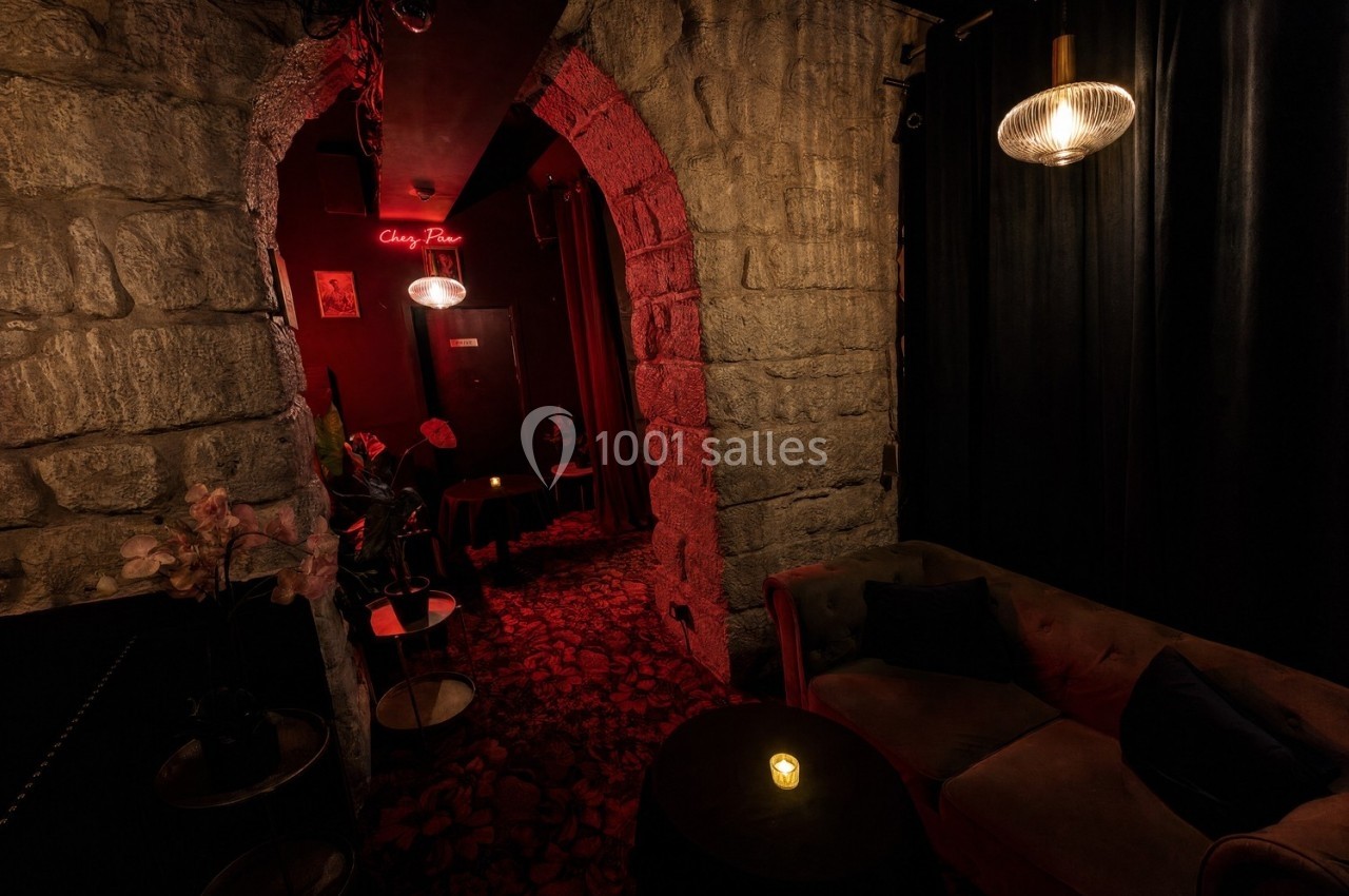 Salon cosy avec canapé, table basse éclairée par une bougie, murs en pierre et lumière tamisée rouge au fond.