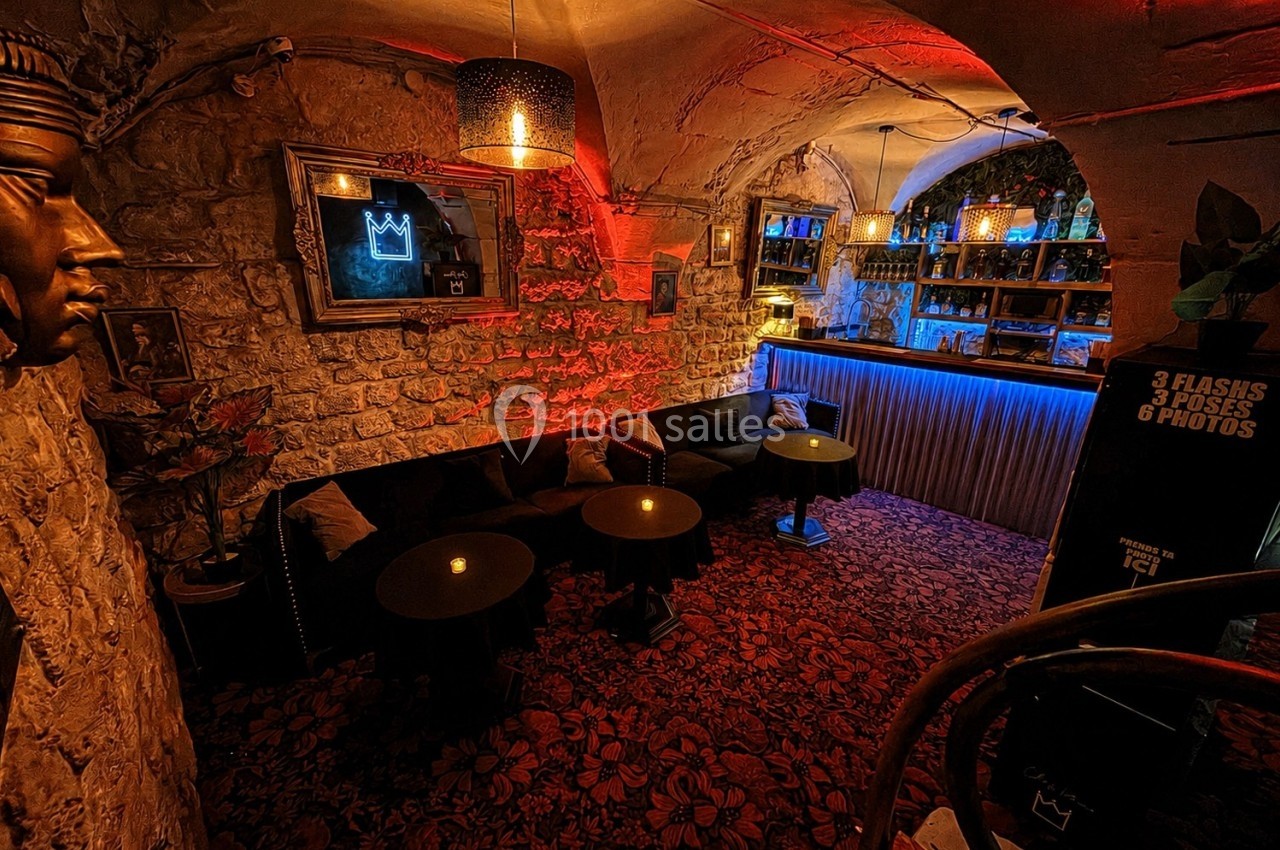 Salle voûtée en pierre avec éclairage tamisé, tables rondes, banquettes et bar illuminé en arrière-plan.
