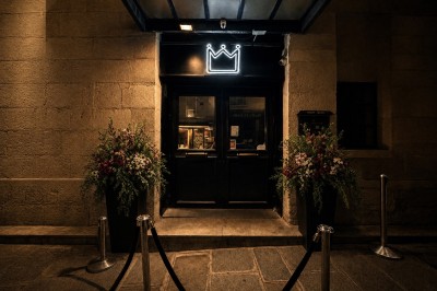 Entrée sombre d'un bâtiment avec un néon en forme de couronne, encadrée par deux grands pots de fleurs.