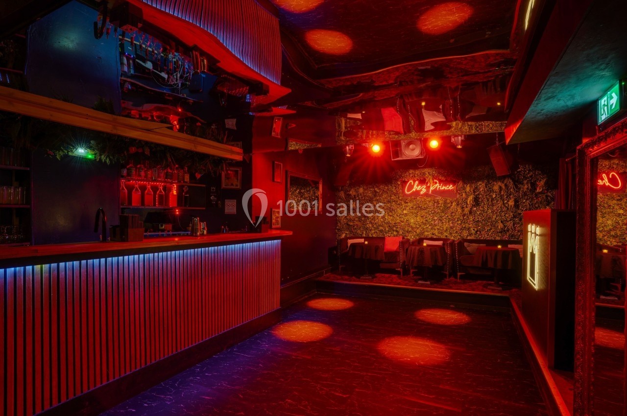 Salle de bar avec éclairage tamisé rouge, comptoir illuminé et tables disposées au fond près d'un mur végétal.