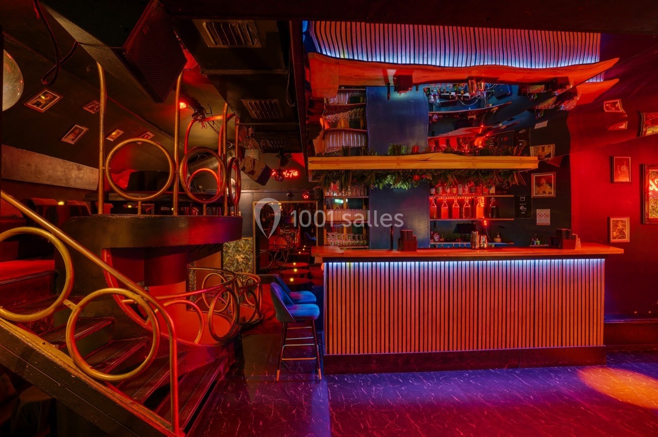 Bar coloré avec éclairage rouge et bleu, comptoir en bois, tabourets et décorations murales éclectiques.