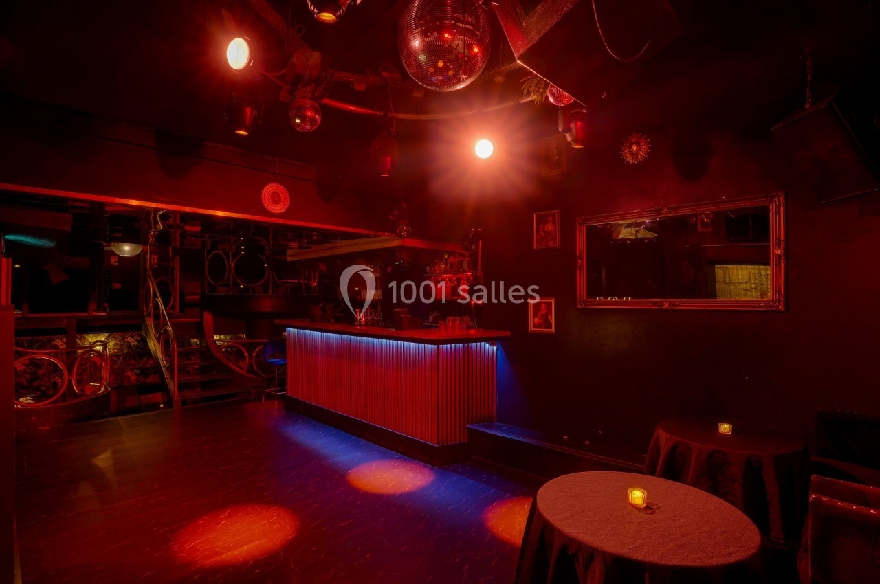 Salle de bar sombre avec éclairage rouge, tables rondes, bougies et une piste de danse avec boule à facettes.
