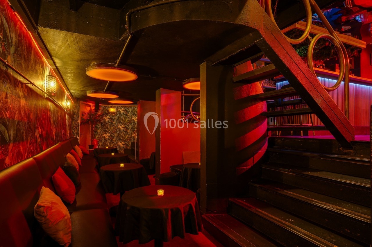 Escalier en colimaçon menant à une salle tamisée avec tables rondes, banquettes et éclairage rouge chaleureux.