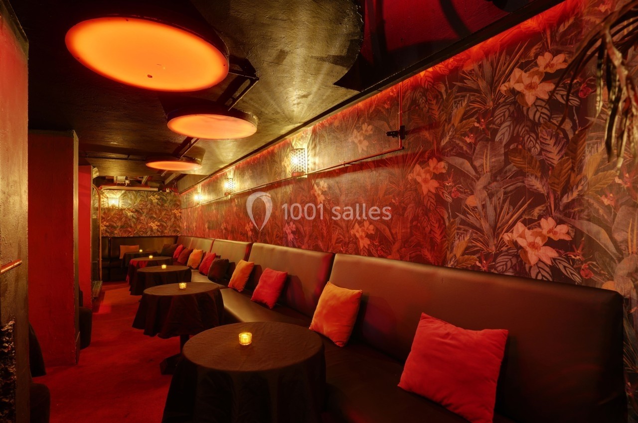 Salle lounge avec banquettes noires, coussins colorés, tables basses et éclairage tamisé rouge, décor tropical aux murs.