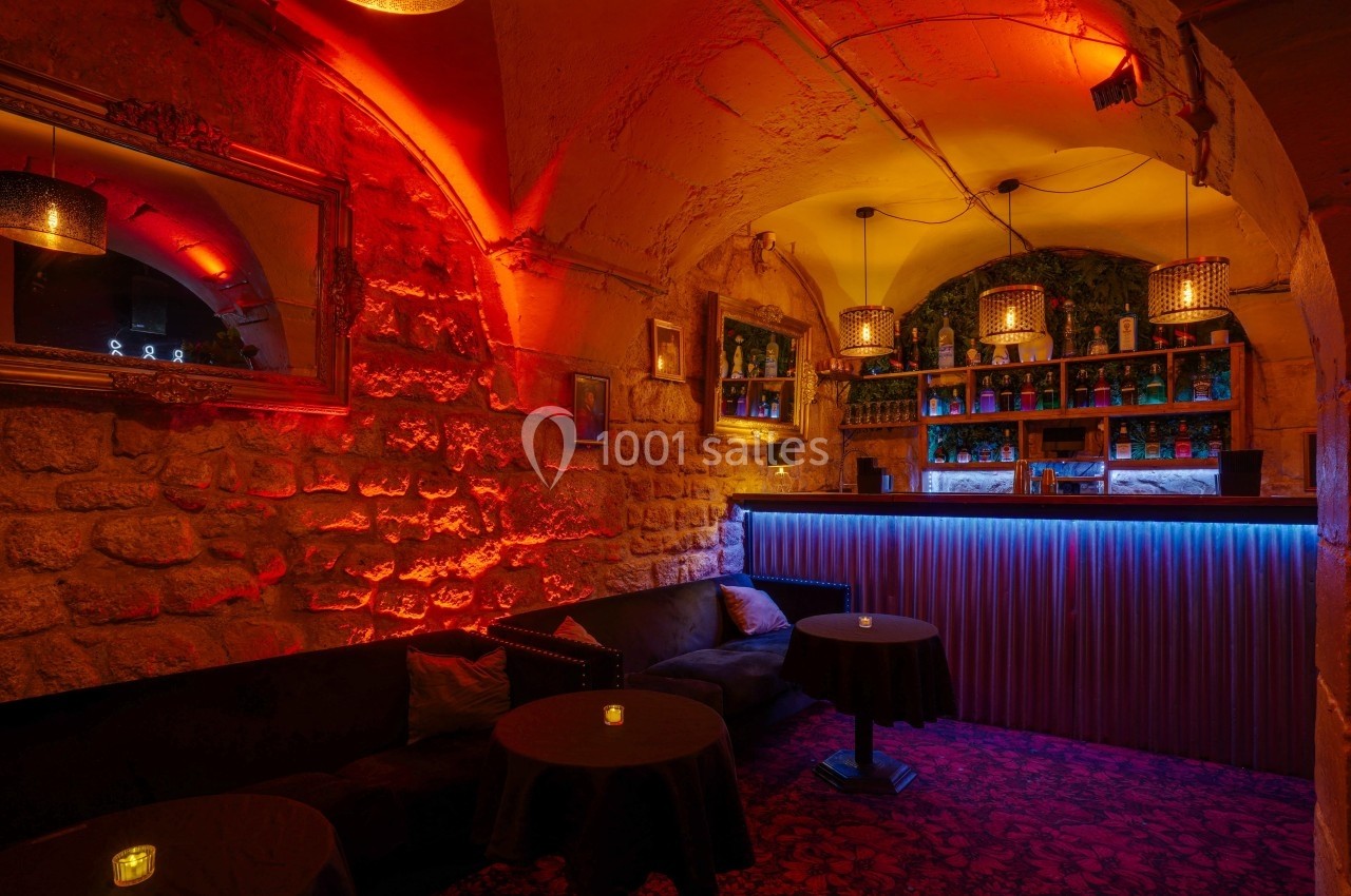Salle de bar voûtée avec murs en pierre, éclairage tamisé rouge et bleu, tables basses et banquettes noires.