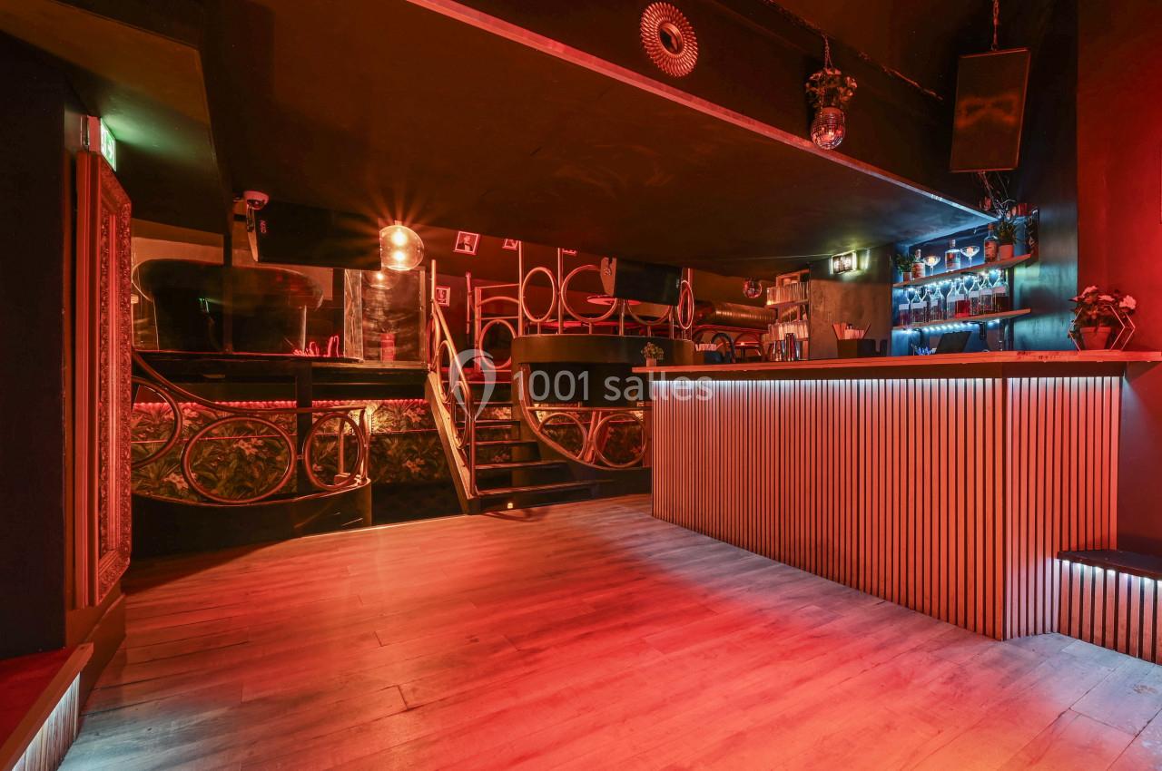 Intérieur d'un bar avec un comptoir éclairé, un escalier menant à une mezzanine et une ambiance lumineuse tamisée.