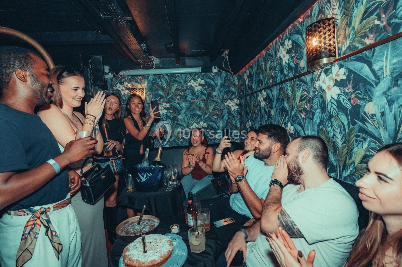 Groupe de personnes célébrant dans un bar décoré de motifs tropicaux, applaudissant autour de gâteaux.