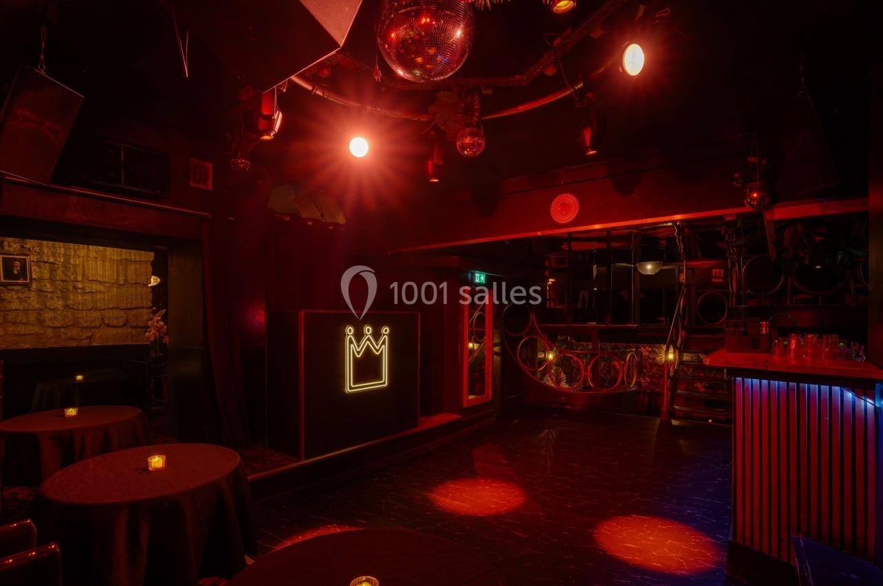 Salle de club sombre avec éclairage rouge, tables rondes, bar éclairé et enseigne lumineuse en forme de couronne.