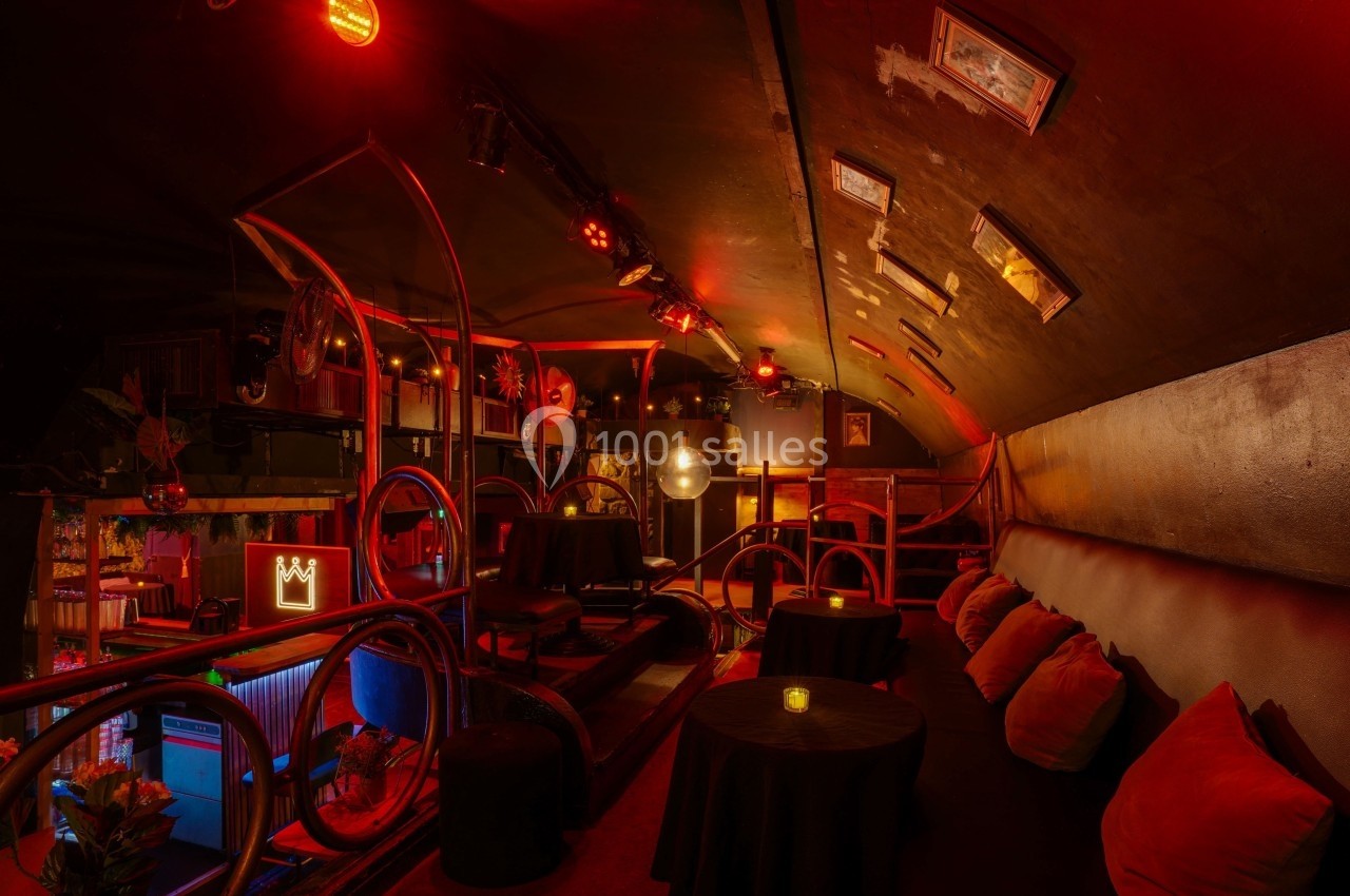 Salle intimiste avec éclairage rouge tamisé, tables rondes, banquettes avec coussins et décorations murales.