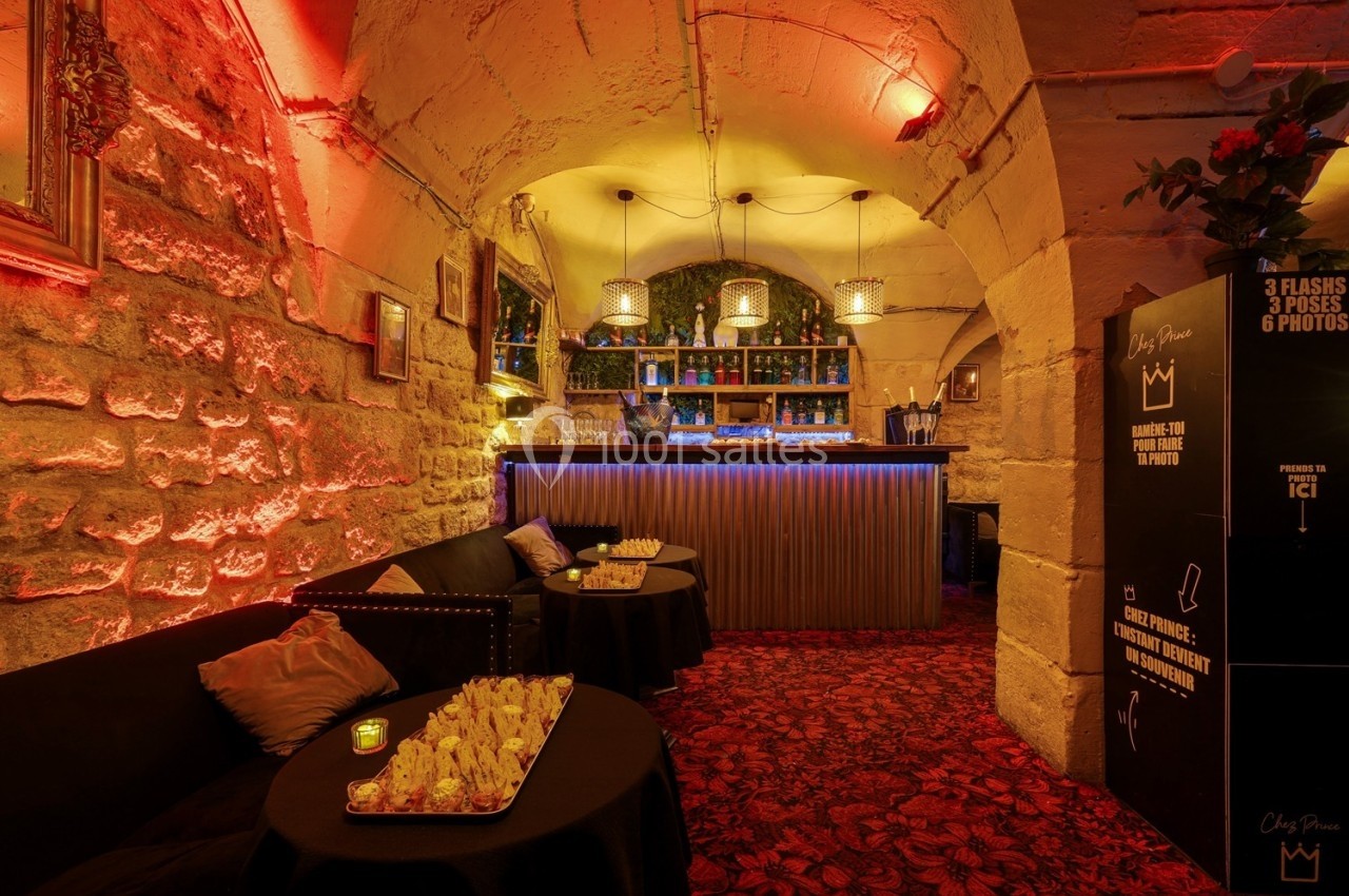 Salle voûtée en pierre avec éclairage chaleureux, bar illuminé, tables garnies et ambiance cosy.