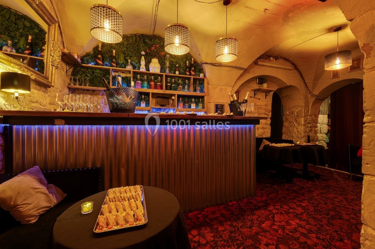 Bar avec comptoir éclairé, étagères garnies de bouteilles, tables dressées et ambiance chaleureuse dans une cave voûtée.