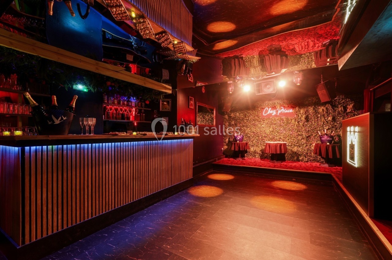 Salle de bar avec comptoir éclairé, piste de danse vide et décorations murales végétales sous une lumière tamisée.